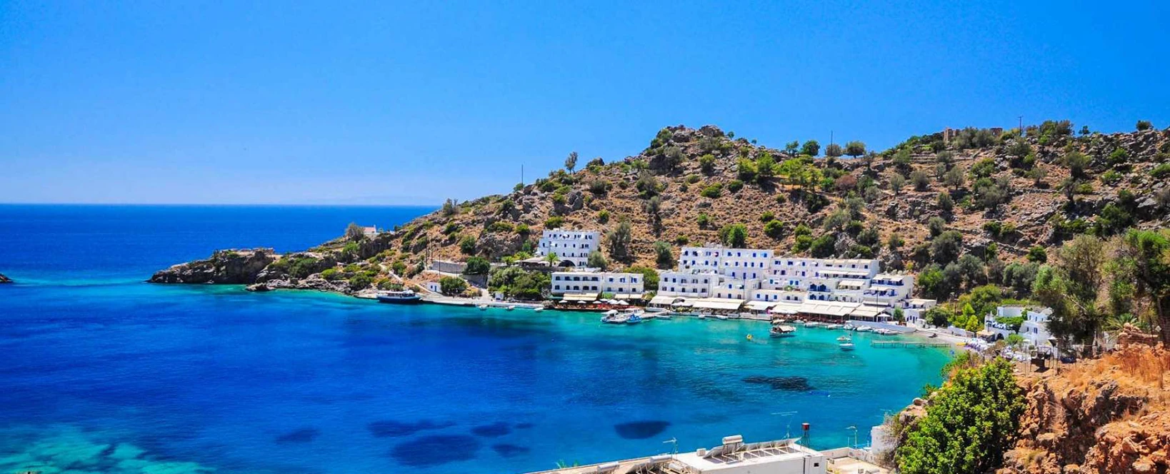 Apa albastra si turcoaz in Loutro, Creta, cu flori roz in prim-plan si case albe traditionale